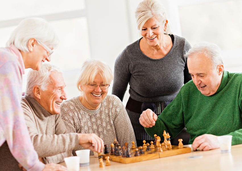 Five seniors who are residents play chess and share laughter as the light shines through the windows on a sunny afternoon.
