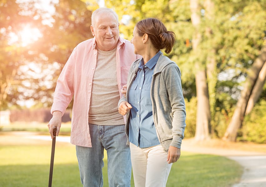 A Culpepper Place employee assists an out of breath man who is walking with a cane outside in the evening sunlight.