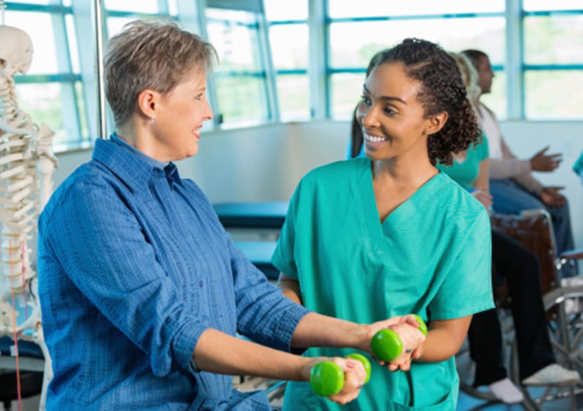 A patient with limited mobility extends free weights out in front of herself while a physical therapist helps them.