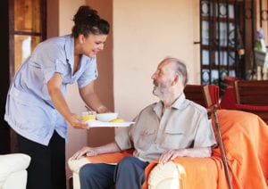 A Culpepper Place employee brings nutritious food and drink on a tray to a resident who is sitting in an armchair.
