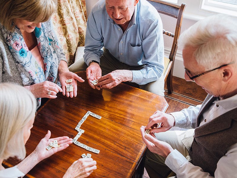 Four assisted living residents are playing dominos together in a recreation room where board games are available.
