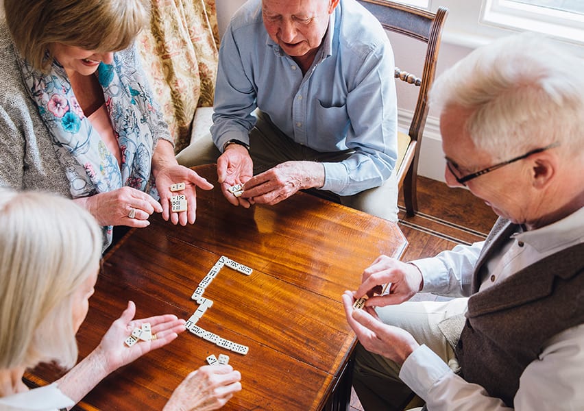 Four assisted living residents are playing dominos together in a recreation room where board games are available.