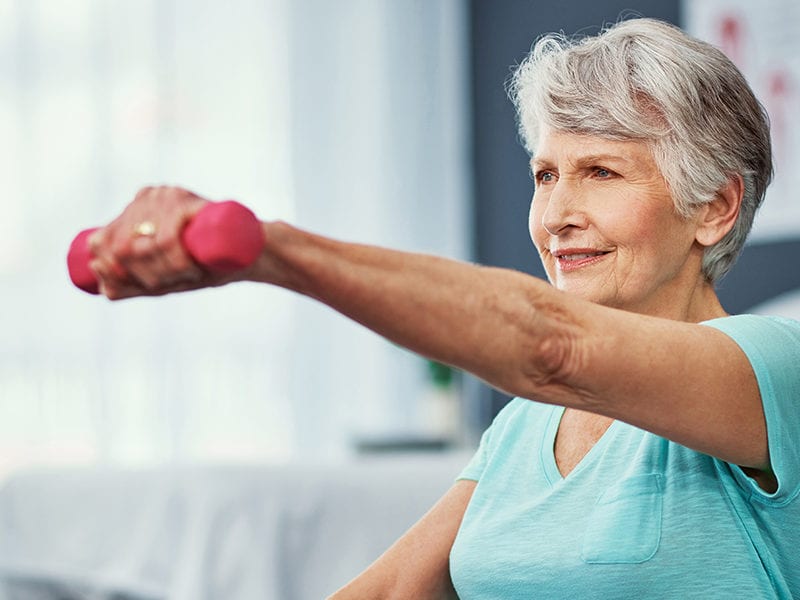 A fit elderly woman holds an exercise pose meant to build strength while extending her arm in front of her with a free weight.