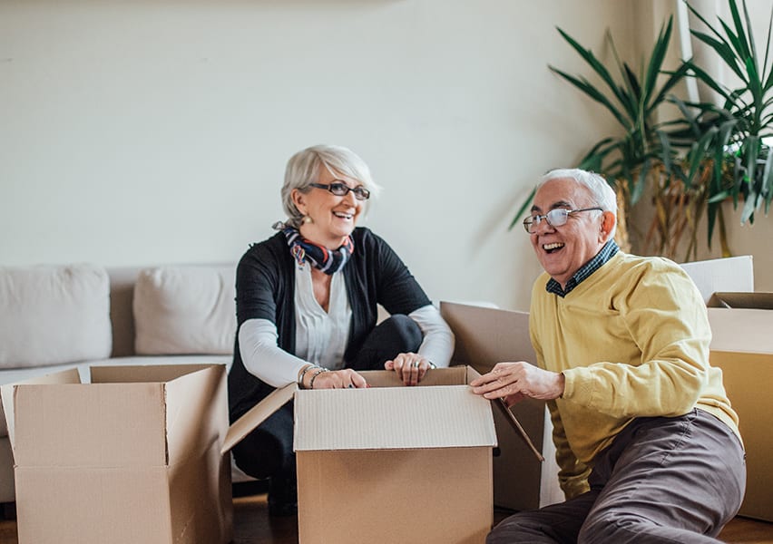 Two seniors moving into their private assisted living apartment unpack and have a good time as they smile from the floor.