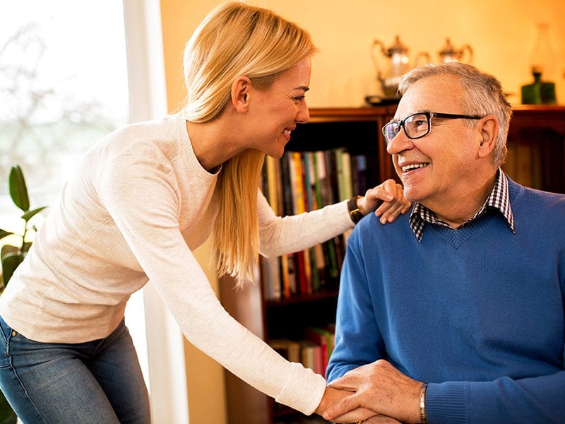 A happy male resident wearing glasses pats one hand of a caregiver who has her other hand on his shoulder.