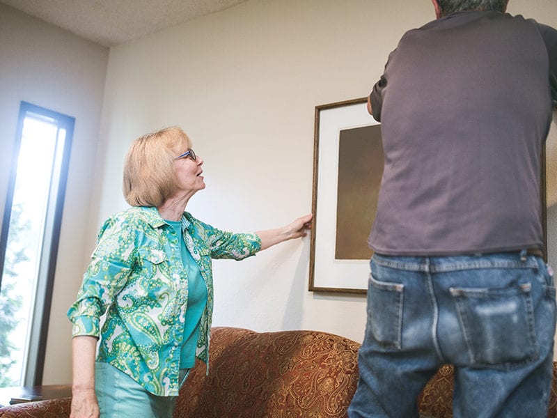 A man helps a senior woman hang artwork on the wall of her assisted living apartment.